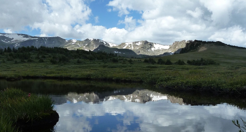 a rocky and snowing mountain ridge is reflected in the still waters of a pond